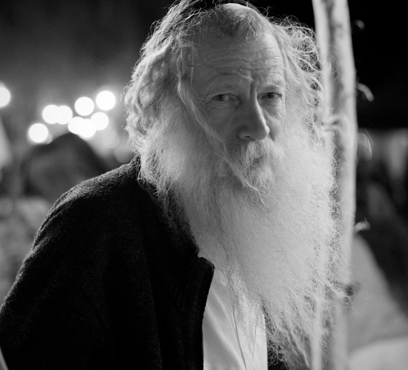 An elderly man with a long white beard looks over his shoulder at the camera, festival lights behind him, Arava Desert