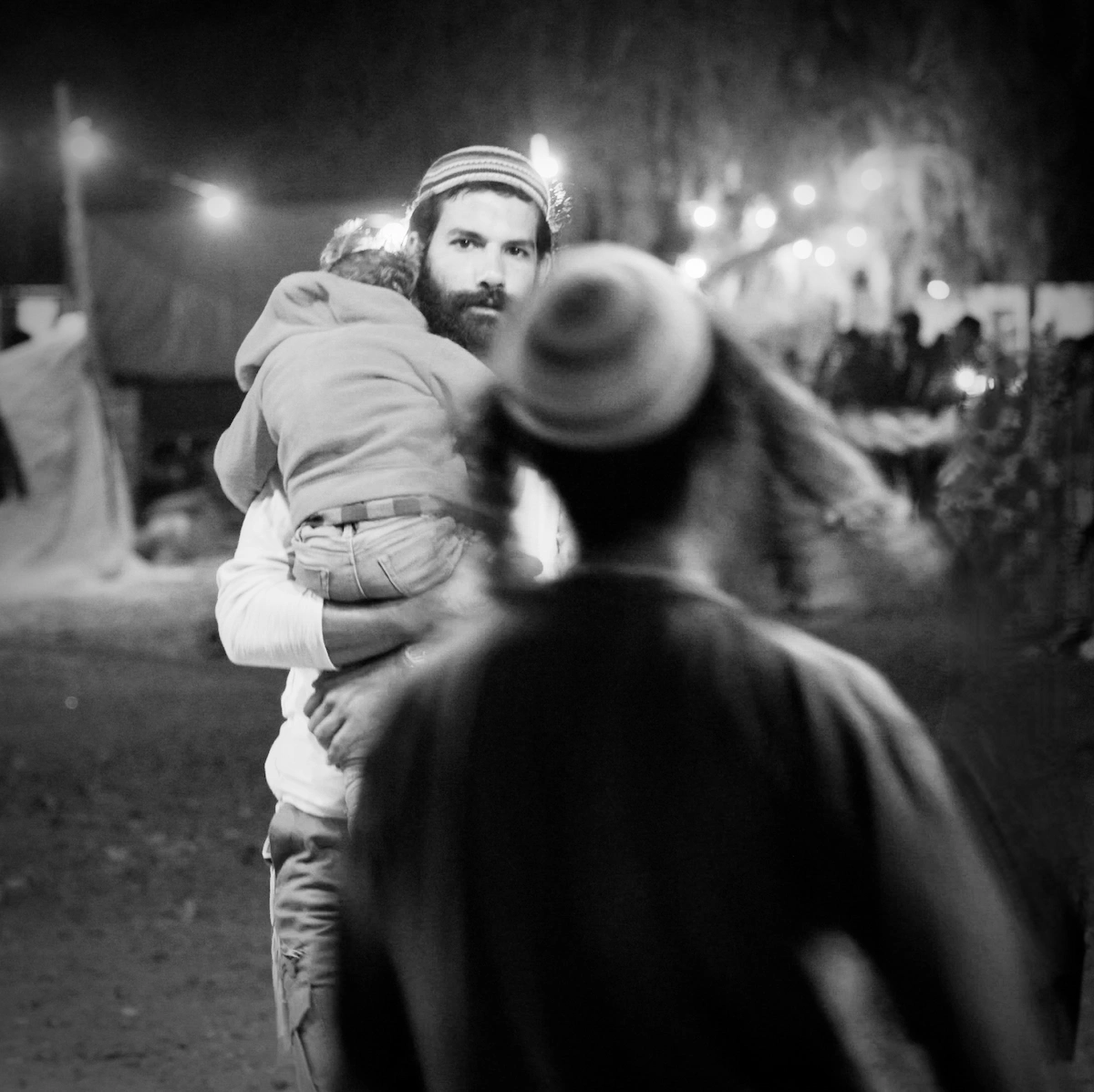 A bearded father carries his sleeping child through a night festival crowd, string lights behind him, Arava Desert