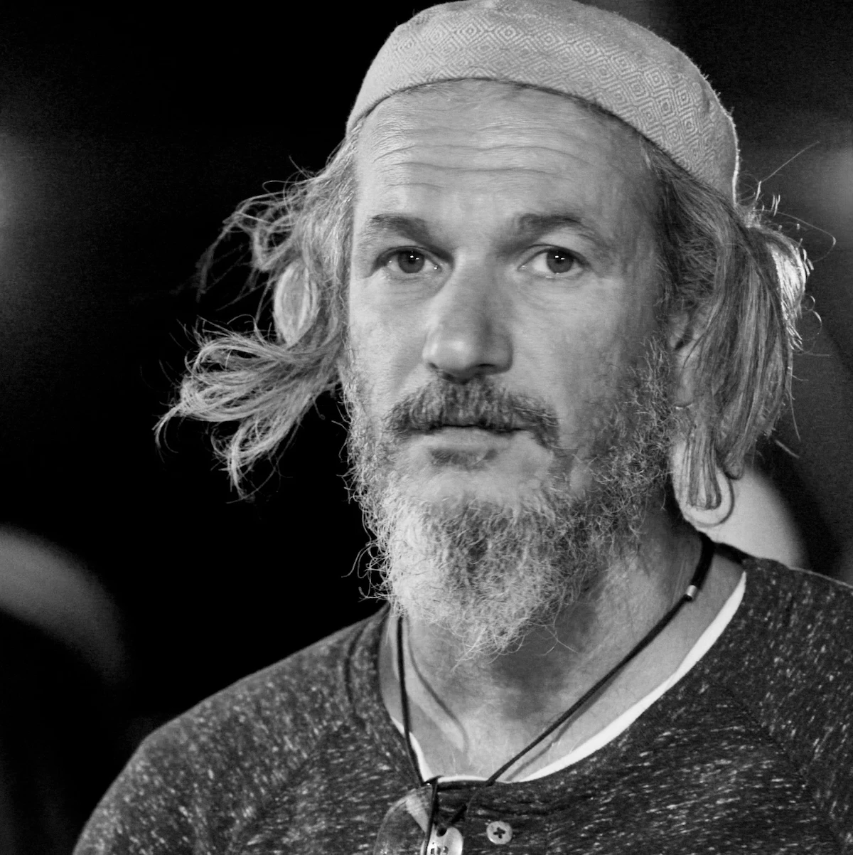 A man in a woven cap, hair lifted by the wind, gazes just past the camera at night, Arava Desert