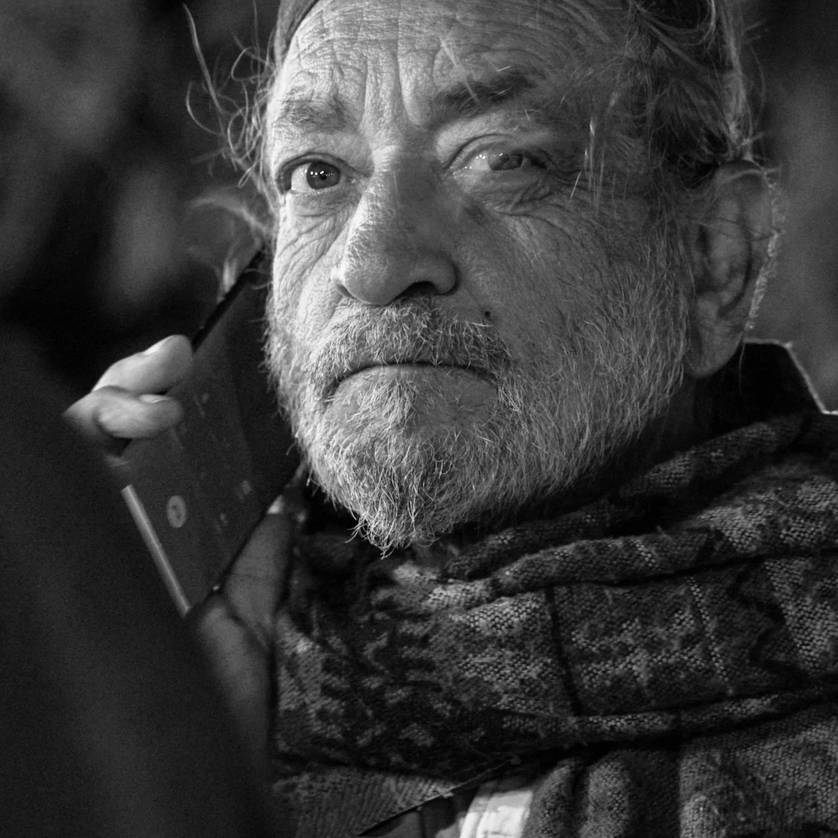 An older man with a deeply lined face listens on a phone, a patterned scarf at his neck, at a night festival in the Arava Desert