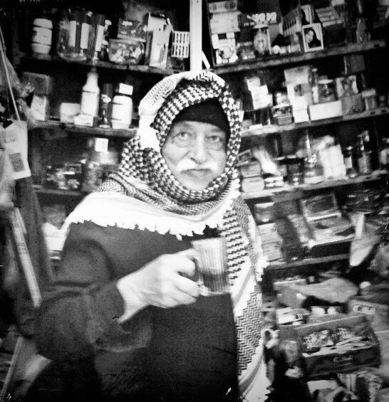 A shopkeeper in a keffiyeh holding a cup, surrounded by shelves of goods, Old City, Jerusalem