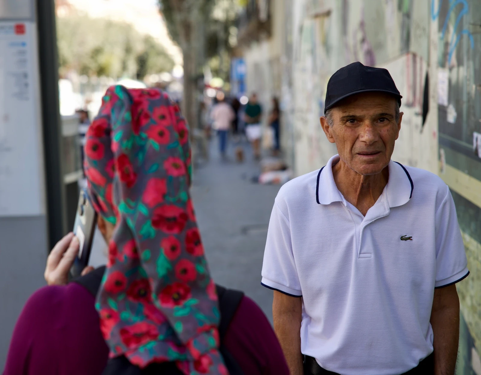 Older man in a black cap and white polo shirt beside a graffiti-covered wall, a woman in a floral headscarf in the foreground, Jerusalem