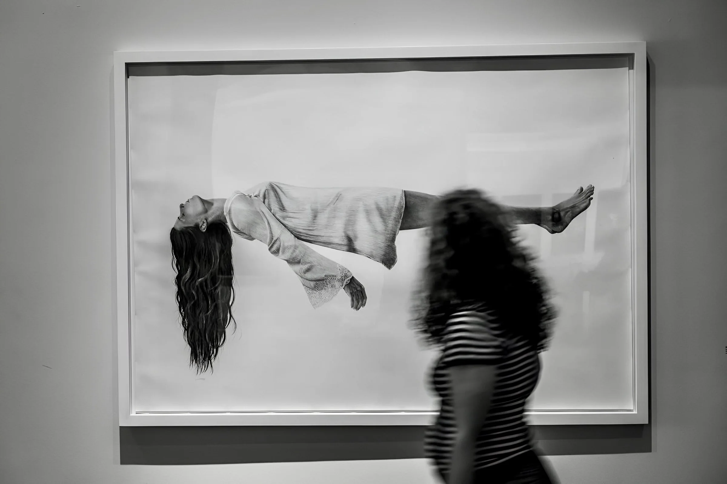 A blurred visitor in a striped shirt walks past a framed drawing of a woman floating horizontally, Israel Museum, Jerusalem