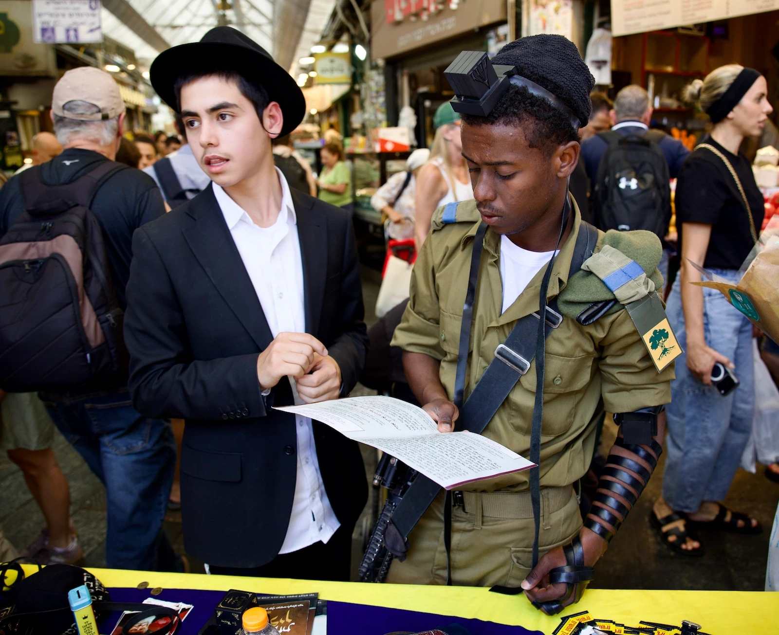 An IDF soldier wearing tefillin reads from a prayer sheet beside a young man in a black hat at a market stall, Jerusalem